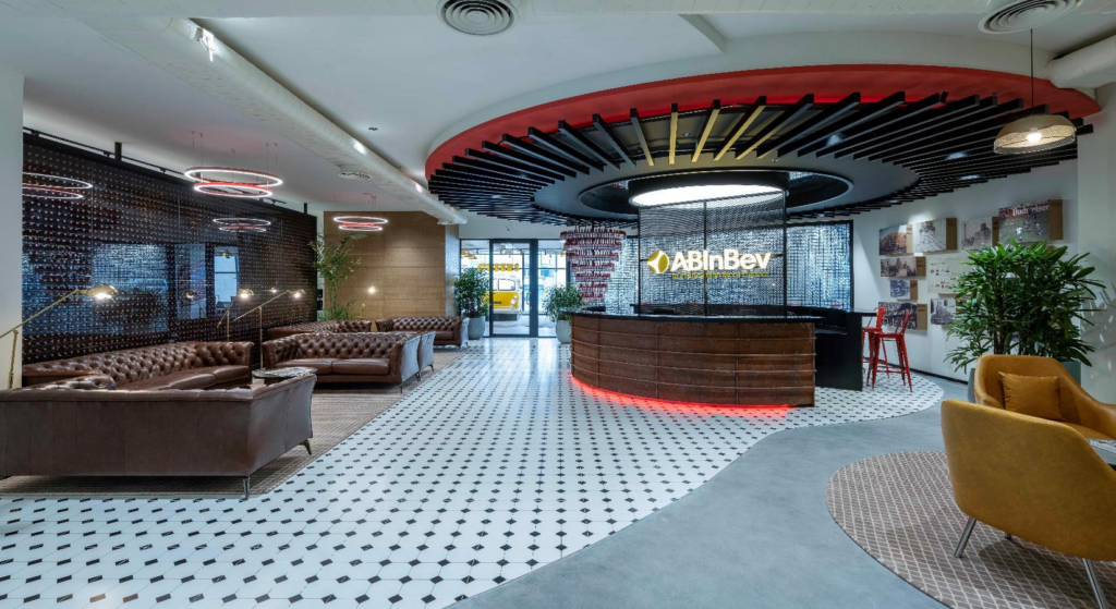 Main reception: curved leather-clad desk with red glow at base, backlit AB InBev mark on perforated screen, black-and-white checkerboard floor, chesterfield lounge.