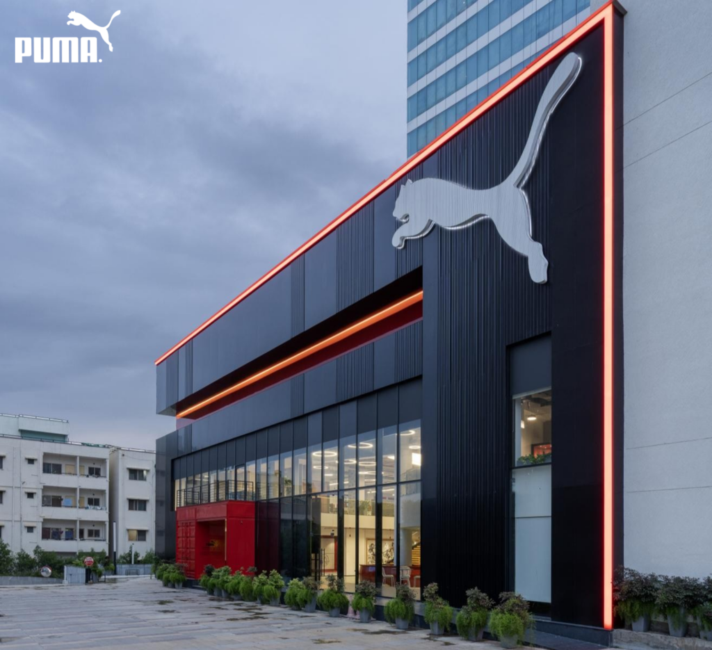 Building exterior at dusk: dark vertical louvered cladding, continuous orange neon brand stripe, illuminated Puma cat on facade.