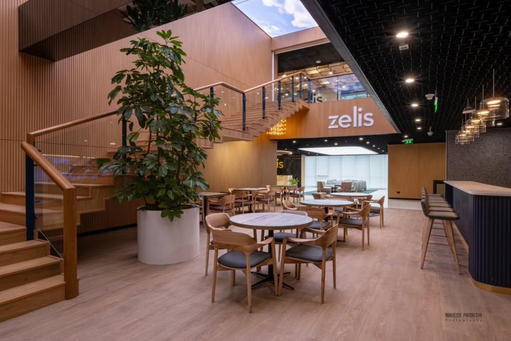 Double-height café with timber floating staircase, wood-slatted ceiling, backlit Zelis brand mark on feature wall, mid-century timber chairs at round tables, soft pendant lighting.